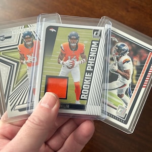 May include: Three football trading cards in protective sleeves. The cards feature players in orange and blue uniforms. One card is labeled "ROOKIE PHENOM" and includes a patch. The cards are held in a hand.