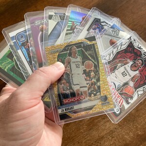 May include: A collection of basketball trading cards featuring Ja Morant of the Memphis Grizzlies, held in protective plastic sleeves. The cards display various designs and colors, including gold and red accents. The word "Monopoly" is visible on one card.