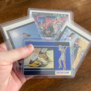 May include: A collection of basketball trading cards in protective sleeves. The cards feature images of basketball players, including Caitlin Clark, and the text "PANINI CHRONICLED - STATE OF THE ART". The cards are displayed on a wooden surface.