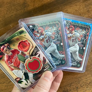 May include: Three baseball trading cards featuring Mike Trout of the Los Angeles Angels. The cards are in protective plastic sleeves. The cards have a Christmas theme with red and green colors. The text on the cards reads "Mike Trout" and "Angels".