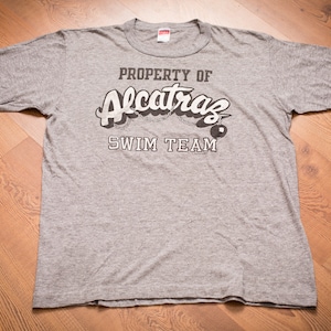 May include: A gray t-shirt with the text "PROPERTY OF ALCATRAZ SWIM TEAM" printed on the front. The text is in a white, stylized font with a black outline. There is a small black and white image of a swim buoy next to the text.