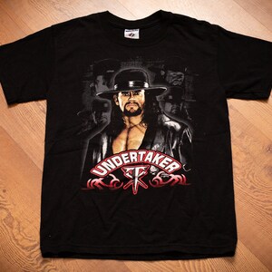 May include: Black t-shirt featuring a graphic of a man in a hat with the word "UNDERTAKER" in red and white. The shirt has a crew neck and short sleeves. The image is a close-up of the shirt.