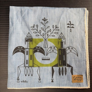May include: Light blue linen handkerchief with a geometric design in black and green. The design features two stylized figures, a tree, and a circular element. The handkerchief has a white border and a small brown label with text.