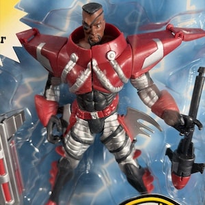 May include: Action figure of a superhero in a clear plastic package. The figure is predominantly red and silver, with black accents. It features a muscular build, an eye patch, and a weapon. The packaging has a yellow and black logo.