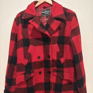 May include: A double-breasted coat featuring a bold red and black buffalo plaid pattern. The coat has a wide collar, long sleeves, and two front flap pockets. A label inside the neck reads White Stag with additional text. The coat is on a hanger.