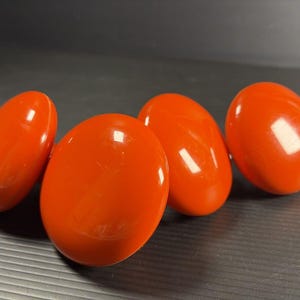 May include: Four round, glossy, bright orange cabinet knobs with a smooth, reflective surface. Each knob features a simple, circular design with a slightly domed top, providing a uniform appearance across the set of four individual hardware pieces.