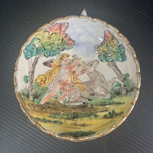 May include: A round, hand-painted ceramic plate with a gold rim. The plate features a pastoral scene with a cherub playing a flute near a deer, trees, and a blue sky. The colors include pink, yellow, green, and blue.