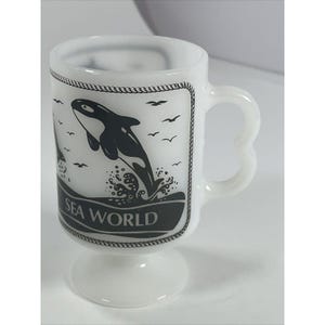 May include: White ceramic Sea World mug with a black and white orca design. The mug features a pedestal base and a heart-shaped handle. The design includes the words "Sea World" and images of birds.