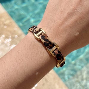 Op de afbeelding: Een armband met een reeks goudkleurige metalen schakels die afwisselen met segmenten van stof met een bruin en zwart luipaardpatroon. Het ontwerp bestaat uit gladde, afgeronde metalen verbindingen verbonden door het materiaal met dierenprint.