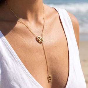 Peut inclure: Un collier lariat doré composé d'une chaîne fine et de deux maillons ovales ornés d'une barre horizontale. Le collier présente une forme en Y, où un maillon ovale sert de connecteur et l'autre de pendentif tombant le long de la chaîne.