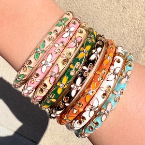 Op de afbeelding: Een stapel van acht dunne metalen armbanden met kleurrijke emaille bloempatronen en kleine ingebedde heldere kristallen. De armbanden zijn uitgevoerd in diverse kleuren zoals lichtroze, groen, zwart, oranje, wit en turquoise, elk met een herhalend bloemmotief.