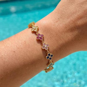 Peut inclure: Un bracelet à chaîne dorée composé d'une série de petites breloques en forme de fleurs à quatre pétales. Chaque fleur est ornée de minuscules pierres précieuses aux couleurs vives, dont le jaune, le rose, le magenta, le bleu et le turquoise.