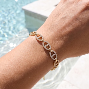 May include: A gold chain bracelet featuring a series of oval-shaped links, each designed with a horizontal bar across the center. The surface of every link is encrusted with small, clear, sparkling stones that catch the light.