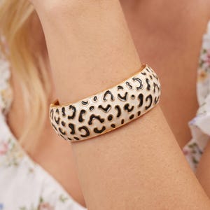 Puede incluir: Una pulsera rígida con bisagra que presenta una superficie de esmalte color crema adornada con un estampado de leopardo negro. La pulsera tiene un borde de metal dorado pulido a lo largo de los lados, lo que le da un aspecto estructurado.