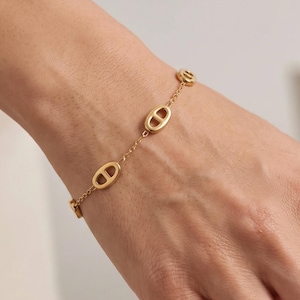 Pode incluir: Uma pulseira de corrente em tom dourado com três elos ovais polidos e espaçados uniformemente, cada um com uma barra horizontal no centro. A pulseira é composta por uma corrente fina e delicada que conecta os elementos ovais mais largos.