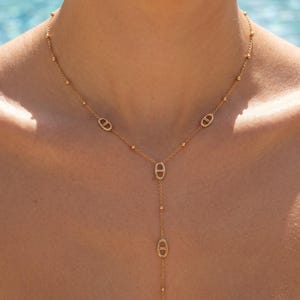 Può includere: Una collana lariat color oro composta da una catenina sottile con piccole perline distanziate uniformemente e cinque maglie ovali, ciascuna con una barra orizzontale centrale. Il design presenta una sezione verticale che termina con una maglia.