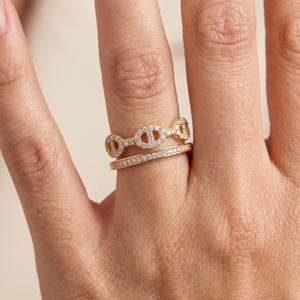 Peut inclure: Un ensemble de deux bagues dorées portées ensemble. La bague supérieure présente un motif de maillons ovales répétés incrustés de petites pierres claires, tandis que la bague inférieure est un anneau fin et simple serti de pierres claires.
