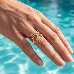 Può includere: Un anello dorato a più fasce composto da tre sottili cerchi collegati tra loro. L'anello è decorato con piccoli ciondoli pendenti a forma di stella, uno dei quali più grande e incastonato con minuscole pietre chiare, accanto a una piccola pietra incastonata.