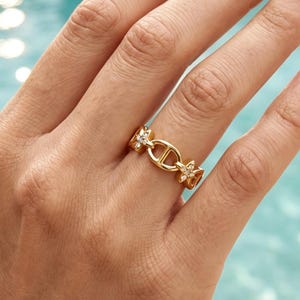 Op de afbeelding: Een goudkleurige ring met een centraal horizontaal ovaal schakelstuk verbonden met twee kleinere bloemvormige accenten, elk ingelegd met kleine heldere steentjes. De ring heeft een gepolijste metalen afwerking en een doorlopende schakelstructuur.