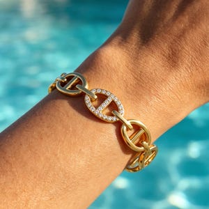 Op de afbeelding: Een gouden schakelarmband met ovale schakels voorzien van een horizontale balk in het midden. Eén van de schakels is aan de buitenrand ingelegd met kleine, heldere, fonkelende steentjes, wat contrasteert met de gladde gouden schakels.