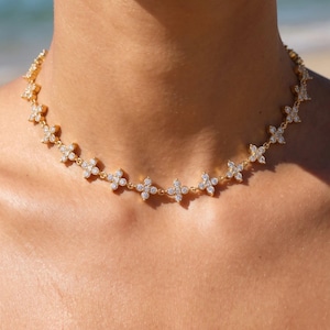 Op de afbeelding: Een goudkleurige choker-ketting bestaande uit een reeks verbonden bloemmotieven met vier blaadjes. Elke bloem is ingelegd met kleine, heldere, fonkelende steentjes. De ketting sluit nauw aan rond de hals en toont een herhalend bloemenpatroon.
