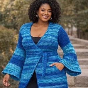 Op de afbeelding: Een blauw, gehaakt wikkelvest met lange mouwen, met een riem en zakken. Het vest heeft een verloop van blauwtinten, van licht naar donker, en heeft wijde klokmouwen. De vrouw draagt er een zwarte top onder.