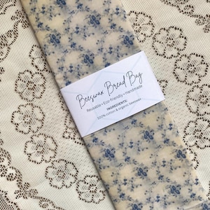 May include: A reusable beeswax bread bag with a cream background and blue floral pattern. The bag is labeled "Beeswax Bread Bag" and lists ingredients as 100% cotton and organic beeswax. The bag is eco-friendly and handmade.