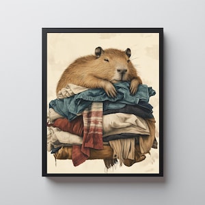 May include: A framed art print featuring a capybara resting on a pile of folded clothes. The capybara has brown fur and is lying down with its eyes closed. The clothes are various colors, including blue, red, and white.