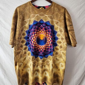 May include: A short-sleeve t-shirt featuring a brown tie-dye pattern with a central geometric mandala design in shades of blue, purple, orange, and yellow. The circular graphic is surrounded by a white lattice-like pattern on the brown fabric.