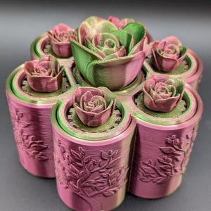 May include: A 3D-printed planter with six compartments, each holding a rose-shaped succulent. The planter is a gradient of pink and green, with a tree and floral design on the sides. The central succulent is larger.