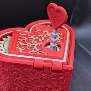 May include: A red heart-shaped music box with a matching heart-shaped key. The box features intricate floral designs and gold accents. The key is inserted into the top of the box, ready to be turned. The box is a decorative item.