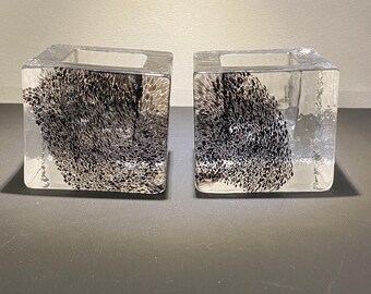 May include: Two clear glass cube candle holders featuring an internal black speckled pattern. Each square vessel has a hollow centre and smooth, transparent sides that reveal the dense, dark, and textured inclusions suspended within the glass.