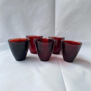 May include: Five dark red glass tumblers with a conical shape are arranged on a white surface. The glasses have a deep, rich color, and the light reflects off their smooth surfaces. The tumblers are likely for serving beverages.