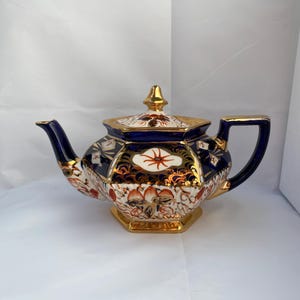 Può includere: Una teiera vintage ornata, con corpo blu navy, finiture dorate e base esagonale. La teiera presenta intricati motivi floreali e geometrici nei toni dell'arancione, bianco e oro. Il coperchio è sormontato da un pomello dorato.