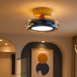May include: A flush mount ceiling light fixture featuring a bright yellow cylindrical base attached to a circular, transparent blue glass shade. The center of the fixture contains a white circular light panel that emits a warm, soft glow.