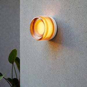 Könnte beinhalten: Moderne Wandleuchte mit kreisförmigem Design. Die Leuchte hat einen transparenten, bernsteinfarbenen Glasschirm und eine weiße Basis, die an einer strukturierten grauen Wand befestigt ist. Das Licht strahlt warm.