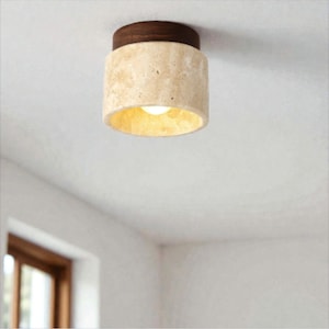 Może przedstawiać: Cylindryczna lampa sufitowa z ciemnobrązowym drewnianym blatem i beżowym, teksturowanym korpusem. Wnętrze lampy jest oświetlone ciepłą, żółtą poświatą. Oprawa jest zamontowana na białym suficie.