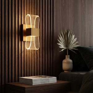 Könnte beinhalten: Eine Wandleuchte mit einem zentralen, rechteckigen, goldfarbenen Sockel und zwei symmetrischen, geschwungenen Schlaufen aus durchscheinendem Acryl, die nach oben und unten ragen und ein warmes Licht durch ein feines Gittermuster abgeben.