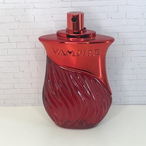 May include: A red glass perfume bottle with a metallic red cap and neck. The word VAMPIRE is embossed on the upper metallic section. The lower portion of the bottle features a textured, wavy glass design in a deep translucent red finish.