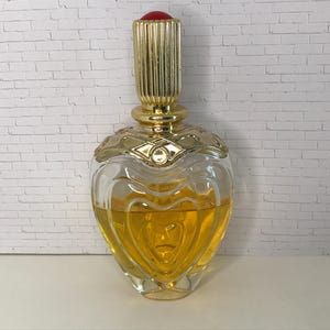 May include: A heart-shaped glass perfume bottle filled with amber liquid. The bottle features a gold-toned metal collar with an embossed pattern and a tall, ribbed gold-toned cylindrical cap topped with a small, smooth red circular accent.