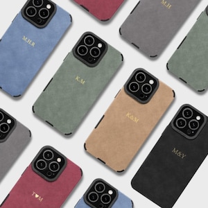 Peut inclure: Une collection de coques pour smartphone dans diverses couleurs, notamment bleu, vert, beige, noir, gris et rouge. Chaque coque présente un contour d'appareil photo noir et des initiales ou symboles dorés gravés comme M.H.R, K.M, K&M, M&Y et T♥M.