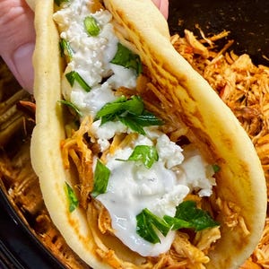 Puede incluir: Primer plano de un sándwich de pan de pita relleno de pollo desmenuzado, queso blanco y una salsa cremosa blanca, adornado con hierbas verdes. El sándwich se sostiene en una mano, con más pollo visible en el fondo.