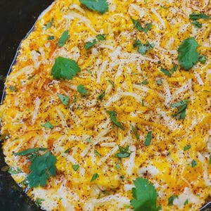 Puede incluir: Primer plano de un plato horneado con una base cremosa blanca, generosamente cubierto con queso rallado amarillo y blanco. Espolvoreado con cilantro verde fresco y pimienta negra, el plato tiene un tono dorado por el horneado.