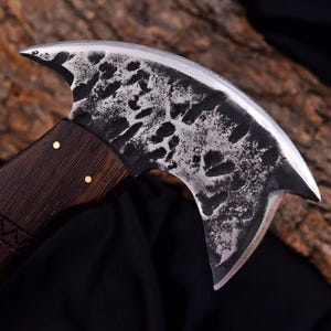 Peut inclure: Une hache artisanale &agrave; lame incurv&eacute;e et manche en bois fonc&eacute;. La t&ecirc;te de la hache pr&eacute;sente un motif textur&eacute; et mouchet&eacute;, fix&eacute;e au manche par des rivets en laiton. La lame est tranchante et incurv&eacute;e, con&ccedil;ue pour couper ou hacher.