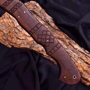Peut inclure: Gros plan sur un manche de couteau en bois fait &agrave; la main. Le manche est de couleur marron fonc&eacute; avec des gravures g&eacute;om&eacute;triques complexes et des accents en laiton. Le grain du bois est visible, ajoutant de la texture. Le manche du couteau est pos&eacute; sur un fond sombre.