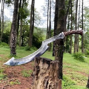 May include: A curved, silver-colored sword with a wooden handle featuring a dotted pattern. The blade has a unique shape with a pointed tip and a curved edge. The sword is displayed outdoors, resting on a tree stump, with a forest background.