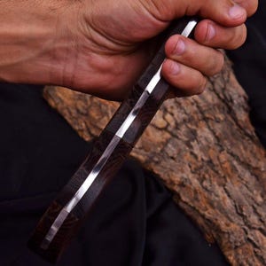 Peut inclure: Gros plan sur un couteau avec un manche en bois fonc&eacute; et une lame argent&eacute;e brillante. Le manche a un aspect textur&eacute; et &agrave; motifs. Le couteau est tenu dans une main, avec un morceau de bois en arri&egrave;re-plan.