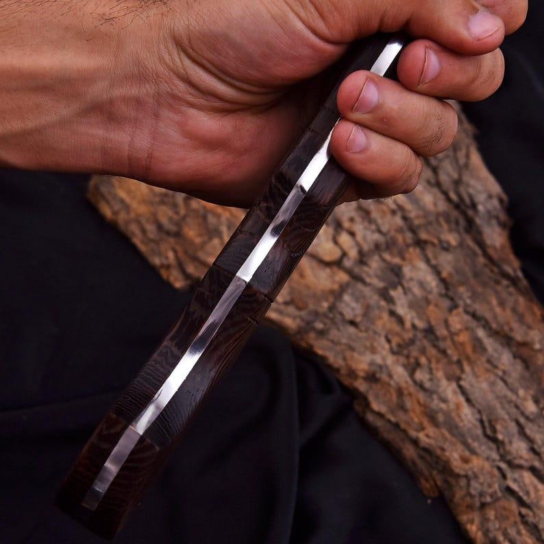 Peut inclure: Gros plan sur un couteau avec un manche en bois fonc&eacute; et une lame argent&eacute;e brillante. Le manche a un aspect textur&eacute; et &agrave; motifs. Le couteau est tenu dans une main, avec un morceau de bois en arri&egrave;re-plan.
