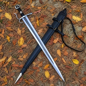 May include: A steel longsword with a black leather-wrapped grip, a silver crossguard with curved quillons, and a central fuller. It is paired with a matching black leather scabbard featuring silver metal fittings and an adjustable leather strap.