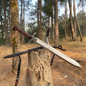 Könnte beinhalten: Ein silbernes Schwert mit braunem Griff und Parierstange, das auf einem Holzstumpf liegt. Ein kleineres Messer mit Ledergriff ist ebenfalls zu sehen. Die Szene spielt sich im Freien mit Bäumen im Hintergrund ab.